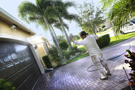 A man pressure washing a garage door on a sunny day, surrounded by palm trees and a landscaped yard. | Beehive Cleaning & Restoration